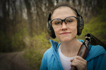 A young woman in shooting gear with hearing and eye protection prepares to shoot a round of sporting clays with an over-under break action shotgun.