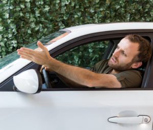 An angry young driver shakes his hand out the window of his white car.